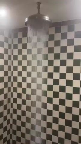 A shower is running in a tiled shower stall. The shower head is a large, round, antique-style fixture with water pouring straight down. The stall is tiled with a checkerboard pattern of dark green and cream-colored square tiles. The lighting is bright, possibly from an overhead light. The water appears to be steaming.