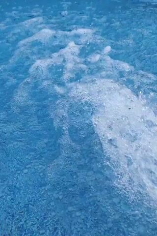 The video shows a close-up, overhead view of bubbling blue water. The water appears to be in a pool or hot tub, with a current of bubbles and white foam flowing from the right side of the frame towards the left. The water is a bright, clear blue, and the bubbles are frothy and white, creating a dynamic and textured surface. There are no people or other objects visible in the frame. The lighting is bright, suggesting an outdoor or well-lit indoor environment. The focus is on the movement and texture of the water.