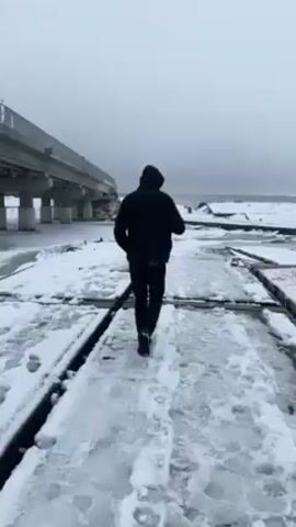A man in black clothes walks along a snowy path towards a partially collapsed bridge. The path is covered in snow and ice, with wooden planks visible underneath. The bridge is damaged, with a large section missing and debris scattered around. The water surrounding the bridge is frozen, and the sky is overcast. The man pauses to observe the scene and then continues walking. The setting appears cold and desolate.