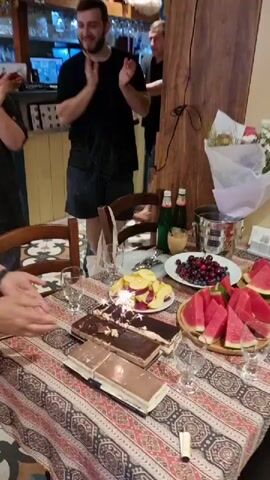 The video shows a group of people gathered around a table laden with food, including watermelon slices and berries. In the foreground, a woman is holding a cake with sparklers that are lit. Several people are clapping and smiling. One man in the background appears to be holding a bottle of alcohol. The setting looks like a restaurant or a home dining area, with visible shelves of bottles and decor on the walls. The overall atmosphere seems celebratory.