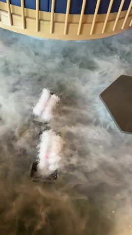 The video shows a restaurant interior with a unique design featuring hexagonal stepping stones over an artificial fog-covered floor. Two fog machines are visible under the fog, emitting plumes. The stepping stones lead towards a backdrop featuring a waterfall design. A round booth with blue cushions and a light wood railing is on the left. A shelving unit is partially visible on the right. People are sitting at a table in the background. The scene is lit with soft ambient lighting.