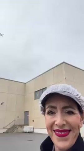 The video features a woman wearing a white hairnet and bright red lipstick, smiling at the camera.  Behind her is a large, beige building with a single window, a door, and a set of stairs leading up to the door.  Above the building, an airplane is visible in the cloudy sky.  A street lamp is also visible to the right of the building.  The woman appears to be standing in a parking lot or similar paved area. The camera remains focused on the woman as the plane flies above her.