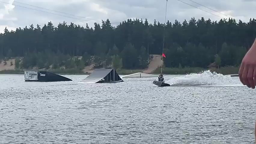 A person is wakeboarding on a lake. The wakeboarder is wearing a dark shirt and shorts and is being pulled by a cable. There are several ramps and obstacles in the water, including a large inflatable water park. The background shows a treeline and a cloudy sky. The video is shot from a slightly elevated perspective, with a wooden dock in the foreground.