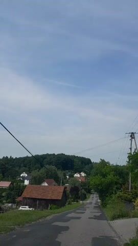 This video captures a scenic drive through a rural Polish village. The camera is mounted to a moving vehicle, giving a first-person perspective of the journey. 

The primary subjects are the picturesque houses and natural landscape. Traditional-looking houses with red-tiled roofs are interspersed among lush green trees and rolling hills that form the backdrop of the scene. A dirt road, patched with asphalt in places, winds through the village, suggesting a somewhat remote or less developed area. Overhead power lines are visible, indicating the presence of electricity in the community.

The activity taking place is a drive through the village, likely for leisure or travel. The time of day appears to be late morning or early afternoon, with the sun casting a soft light. The weather seems pleasant, with a clear blue sky dotted with a few wispy clouds. The overall mood is tranquil and idyllic, conveying a sense of peace and rural charm.

There is no visible text in the video. The location, Michałowice, Poland, provides a specific geographical context to the visual elements. The combination of the natural beauty and the quaint village architecture creates a compelling portrait of Polish countryside life.