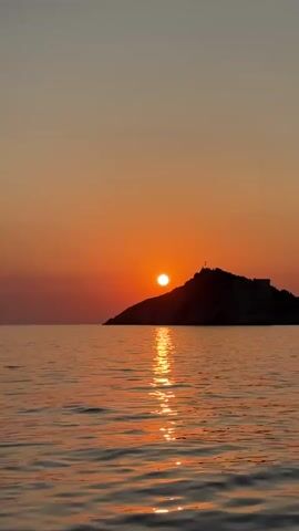 The video shows a serene sunset over a body of water. The sun, a bright orange orb, is setting behind a silhouetted island or landmass. The sky is a gradient of warm oranges and soft purples, transitioning to a muted blue at the top. The sun's reflection creates a shimmering, golden path across the rippling water. The water itself appears calm, with gentle waves. The island is dark and indistinct, with what appears to be a structure or cross at its summit. The overall atmosphere is peaceful and tranquil. There are no people or animals visible in the video. The lighting suggests it is either sunrise or sunset, with the sun positioned low on the horizon.