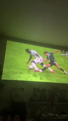 A projection of a soccer game is displayed on a wall. Two players are on the ground, one on his back and the other on his hands and knees. A third player is running towards the left of the screen. The player on his hands and knees is looking intently at the player on his back. Another player's arm is visible on the right side of the screen. The projected image is vibrant green with white lines indicating the field. The room is dark, with the projected image casting a green hue.
