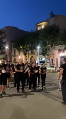 A group of people wearing black t-shirts with a green and red emblem are marching down a street at dusk. They are all holding white objects, possibly drumsticks or batons, and appear to be part of a marching band or parade. The background shows buildings, trees, and streetlights, indicating an urban setting. The sky is dark blue, suggesting it is nighttime or late evening. One person at the front of the group is partially visible from behind, holding drumsticks and a drum. The group is moving from left to right across the frame.