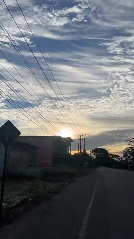 This video is a first-person perspective of a person walking down a paved road at sunrise. The sun is visible on the horizon, casting a warm glow. The sky is filled with scattered clouds. On the left side of the road, there is a building with a red sign and some construction materials. There are also power lines running along the left side of the road. On the right side of the road, there are trees and some tents or market stalls set up. The road appears to be in a rural or suburban area.