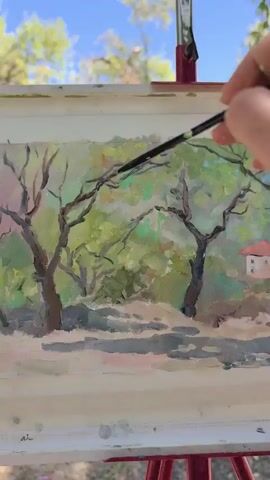 A hand is holding a paintbrush and is in the process of painting a landscape on a canvas. The landscape appears to be a natural scene with trees and a small building with a red roof. The sky is visible in the background, with clouds. The painting is on an easel, which is set up outdoors. The scene is well-lit, suggesting it is daytime.