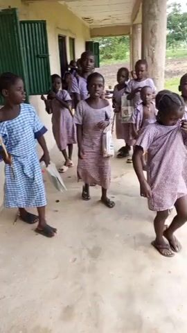 A group of children, mostly girls, are standing on a concrete walkway in front of a building. The children are wearing matching checkered dresses in light purple and white. Some of the children are holding sticks, and one girl in the foreground is holding a white plastic bag. The building appears to be a school, with green shutters on the windows. The walkway is wide and has a concrete floor and a covered roof supported by pillars. The background shows some vegetation and trees. The children are looking in various directions, and some appear to be interacting with each other. The overall scene suggests an outdoor activity or a break time at a school.