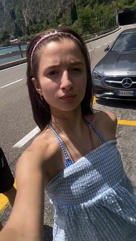 A woman with long dark hair, wearing a blue and white checkered bikini and a light blue and white checkered towel, and a pink headband is taking a selfie video. She is outdoors, with a person in a white t-shirt and black shorts partially visible in the background to her left. Behind them is a paved area with yellow lines, a guardrail, a body of water with boats, and a steep, rocky mountainside covered in vegetation. A car, possibly a grey Mercedes, is parked on the right side of the frame. The video also shows the woman's arm and shoulder, as well as part of the towel. The sun is shining, casting shadows and highlighting the scene.