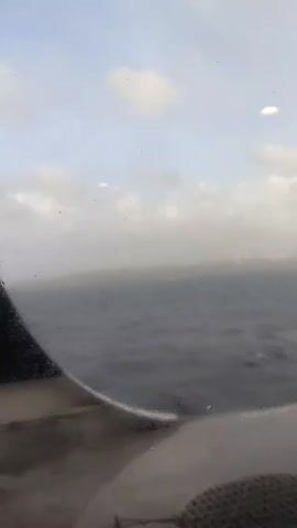 Okay, here's a detailed description of the video:

**Scene:** The video shows a view from the window of what appears to be a boat or ship. The primary focus is on the choppy, dark grey ocean.

**Environment:** 
*   **Weather:** The weather appears overcast and potentially stormy. There is a lot of mist or spray obscuring the view.
*   **Horizon:** A vague, indistinct coastline is visible in the distance, but it is heavily obscured by fog or mist.

**Objects:**
*   **Window:** The video is shot through a window that has some water droplets on it.
*   **Boat/Ship Structure:** Parts of the boat or ship's structure are visible around the window frame, including what seems to be metal and a section with a mesh-like pattern.

**Action:**
* The vessel is moving through the water. The waves are visibly breaking. 
* There are a few whitecaps or pieces of debris visible on the surface of the water.