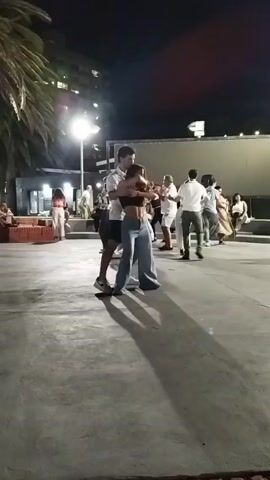 At night, a group of people gather on a paved outdoor space, illuminated by streetlights, to dance. The focal point is a couple dancing in the foreground. The man is wearing a white shirt and dark shorts, while the woman is in a dark top and wide-legged jeans. Other couples can be seen dancing in the background, some wearing white attire. The setting includes a palm tree on the left and a building visible in the distance. Some people are seated on the side, observing the dancers.