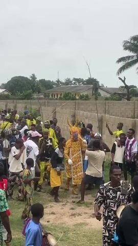 A group of people are gathered outdoors on a dirt path, with a low brick wall and some buildings in the background. The sky is overcast. In the center of the crowd, a person is wearing a vibrant yellow costume with intricate patterns and a mask that resembles an animal with horns. They are being followed and surrounded by other people, some of whom are holding sticks. Some individuals are wearing colorful patterned clothing, while others are in casual attire like t-shirts and shorts. A few people appear to be watching or taking photos with their phones. The overall atmosphere seems to be celebratory or part of a procession.