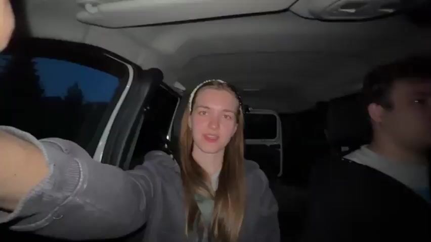 A woman is in the passenger seat of a van or other large vehicle, taking a selfie video in low light. A man is driving, visible only in the periphery. The woman is wearing a gray hooded jacket and a headband. The vehicle appears to be moving. The video is shaky. Loud music is playing.