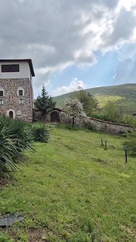 The video shows a building with a red tile roof in a rural setting. The building has a white exterior with dark wooden balconies and a cross above the door. A man in a camouflage jacket walks through the courtyard, which has grass, trees, and a stone wall. There is a car parked in the courtyard. In the background, there is a green mountain range under a cloudy sky. The video also captures the sound of a dog barking.