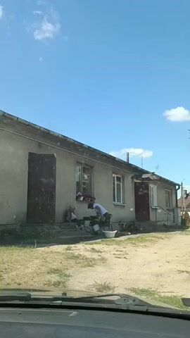 This video captures a scene outside a modest, light-colored house under a bright, clear blue sky with a few scattered white clouds, suggesting a pleasant, sunny day, likely in the morning or early afternoon. The house has a weathered appearance, with a large, dark wooden door on the left and another similar door to the right of the entrance, flanked by windows. A porch area extends from the right side of the house.

In front of the house, two individuals are engaged in activity. An older person, possibly a woman, is seated on the steps to the left. To the right of the seated person, another individual, dressed in a white shirt and dark pants, appears to be tending to something on the ground, possibly a plant or a small animal, with a white basin nearby. This person then moves away from the immediate area in front of the house.

The ground in front of the house is a mixture of dry, patchy grass and sandy dirt, indicating a rural or semi-rural setting. The overall impression is one of everyday life unfolding in a quiet, possibly village environment. There is no visible text in the video. The activity suggests a domestic or personal task being performed outdoors. The mood appears calm and unhurried, typical of a peaceful daytime setting.