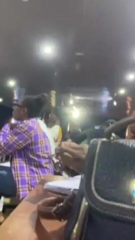 The video shows a partially obscured view of a crowded interior space, possibly a restaurant or meeting hall. Multiple people are visible, some seated at tables. A woman in a purple and white plaid shirt is seated to the left, with other individuals in the background. In the foreground is a textured black handbag with brown leather detailing, positioned on a table. One person's hands can be seen as they manipulate a pair of eyeglasses. The lighting in the room is provided by recessed lights in the ceiling, creating a diffuse illumination. The overall atmosphere suggests a social gathering or busy public space.