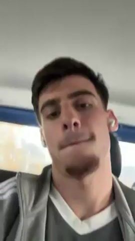A man wearing a gray and white zip-up jacket and white earbuds films himself sitting in a car. He looks into the camera then turns the camera to the left, showing a second man in the car. The second man is Black and is wearing a black baseball cap and black hoodie. He is sleeping in the passenger seat of the car. The first man looks into the camera again. The interior of the car is gray and blue. The view outside the window is blurry, suggesting the car is moving.