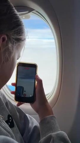 A young woman is shown in profile, seated on an airplane and looking out the window. She holds a smartphone in her right hand, aiming it towards the window to capture a video. The phone screen displays a recording interface, showing the airplane wing and clouds outside. Her hair is pulled back, and she wears a gray hooded sweatshirt. The environment is the interior of an airplane cabin, with a large oval window framing the view of the sky.