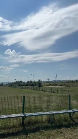The video shows an outdoor scene with a blue sky and white clouds. In the foreground, there is a metal bench with green supports, behind a wire fence with green posts. Beyond the fence, there is a grassy field with several other benches and more fencing. Further in the background, there are utility poles and some trees. To the right, there is a blue portable toilet and a green seesaw. The scene appears to be a park or playground area.