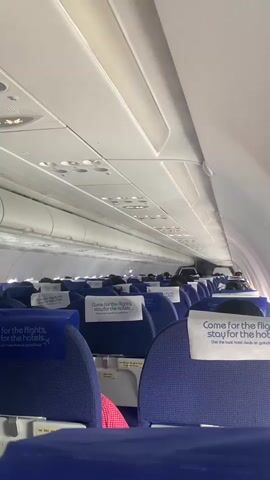 The video shows the interior of an airplane cabin. The seats are mostly blue, and many have white headrest covers that read "Come for the flights, stay for the hotels. Get the best hotel deals on goindigo.in." Passengers are sitting in some of the seats, though many are not visible or are obscured by seatbacks. The ceiling of the plane is white with vents and lights. The cabin appears to be moderately full, though some empty seats are visible. The lighting in the cabin is bright and somewhat diffuse. The camera is still, and the focus is sharp.