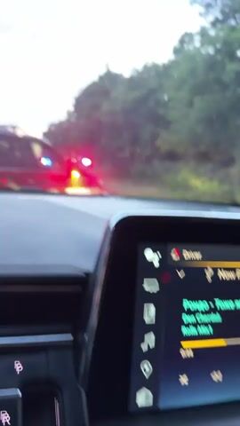 The video shows a view from inside a car looking out the windshield at a traffic scene.  There are several vehicles ahead, including what appear to be police cars with flashing blue and red lights.  The time appears to be dusk or evening, as the sky is overcast with a visible sunset glow.  Trees line the side of the road, and the traffic is either stopped or moving very slowly. The interior of the car is visible in the foreground, including the dashboard and a screen.