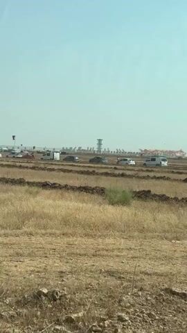 The video captures a dry, open field with rows of tilled earth. In the distance, a line of vehicles, including cars and vans, are parked. Beyond the vehicles, there appears to be an event or gathering with a tall structure resembling a tower and what looks like an aircraft or large inflatable structure with red and orange coloring. The sky is clear and blue, suggesting a sunny day. The overall environment is rural and arid.