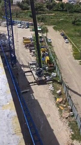 The video shows a construction site in Mirnovskoye rural settlement, Ukraine. The scene is dominated by a large green and grey mobile crane, its boom extended and angled upwards. The crane is positioned on a flatbed truck with outriggers extended for stability. Various construction materials are visible around the crane, including stacks of yellow and blue scaffolding, bundles of lumber, and piles of white bricks. In the background, a blue tower crane stands tall, suggesting ongoing construction of a large structure. Other vehicles, including cars and a white van, are parked in the area, along with some portable buildings. A metal fence borders part of the site, and beyond that, a dirt road leads to a more developed area with buildings and trees. The lighting suggests it is daytime, likely sunny given the bright illumination. The overall impression is a busy construction environment. There is no discernible human activity or emotion visible. The visible text on the crane includes branding and possibly model information, but it is too small to be clearly read. License plates on the back of the crane truck are also visible but unreadable.