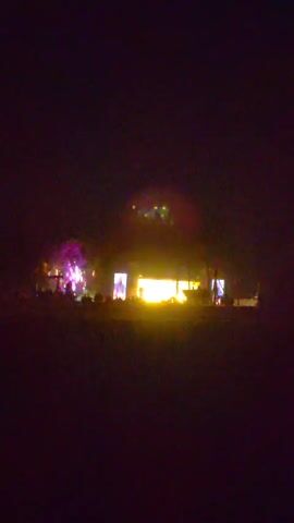 The video shows a nighttime outdoor event with a stage illuminated by bright yellow and pink lights. Several screens are set up on the stage, displaying what appear to be images or videos of people. A crowd is gathered in front of the stage, silhouetted against the lights. Some lights are also visible in the sky above the stage, possibly fireworks or spotlights. The overall atmosphere suggests a concert or festival.
