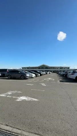 The video begins with a wide shot of a parking lot in front of a modern, elongated building with a glass facade and a peaked roof. The sky is a clear, vibrant blue with a few scattered clouds. Cars are parked in rows in the lot, and the pavement is marked with white lines. The camera pans slightly to the right, revealing more parked cars and streetlights. As the video continues, the camera shifts to a lower angle, showing a side mirror of a vehicle in the foreground. The sun is bright and high in the sky, casting long shadows. The building in the background appears to be an airport terminal, with a control tower visible in the distance. The video concludes with a closer view of several parked cars and the control tower.