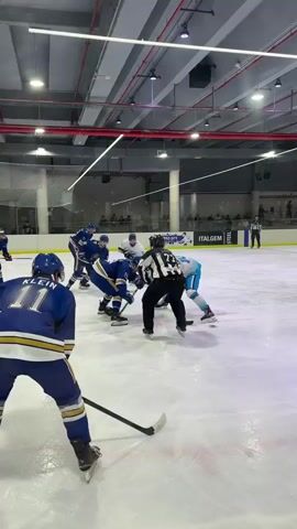 The video shows an ice hockey game. Several players in blue and white uniforms are on the ice. A referee in black and white stripes stands in the center of the ice. Players are skating, holding hockey sticks, and some appear to be engaged in a face-off. The environment is an indoor ice rink with bright lighting and spectators visible in the background. The camera is focused on the action from an angle behind one of the blue-uniformed players. The video captures the initial moments of the game or a face-off.