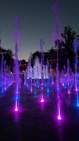 This is a vertical video of a fountain at night. The fountain has multiple streams of water shooting upwards, illuminated by purple and blue lights. The lights create a vibrant, colorful effect on the water and the surrounding pavement. The background is dark, with some trees and buildings visible, suggesting an urban park or public square. The sky is a deep twilight blue. The camera is positioned at ground level, looking up at the fountain, which dominates the frame. There are no people clearly visible in the video. The overall impression is one of a lively and aesthetically pleasing nighttime scene.