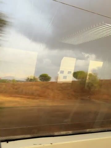 The video shows a view from inside a moving train, looking out the window. The window is wet with raindrops. Outside, a landscape of brown, dry vegetation and hills can be seen. In the distance, there are trees and some buildings. The sky is overcast with dark clouds. Reflections of the train's interior, including what appear to be seats with horizontal lines, are visible on the window. The camera is panning slowly, giving a sense of movement.