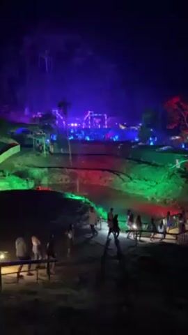 The video shows a night view of a brightly lit outdoor event. A group of people are walking down a path, with some pausing to observe the scene. The event venue, situated on a higher level, is illuminated with blue, purple, and white lights, creating a festive atmosphere. The surrounding landscape is enhanced with green and red lights, and a water feature is colored red and green as well. The sky is a deep blue.