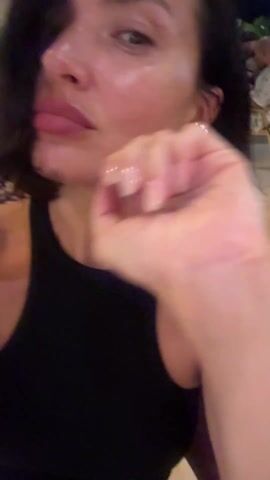 A woman with short, dark hair and gray eyes poses for a selfie. She is wearing a black top and diamond stud earrings. She rests her chin on her right hand. The background appears to be a restaurant patio at night. Other people are seated nearby. The camera blurs at times during the recording.