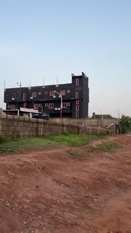 The video shows a black, multi-story building with pink window frames. The building is behind a low, block wall, with patches of green grass and red dirt visible in the foreground. A street light stands in front of the building, painted with white and red stripes. The sky is overcast.