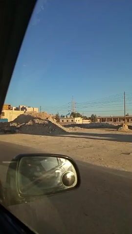 The video captures a street scene from the perspective of a vehicle moving along a road in Sidi Abdelrahman, Egypt, under a clear, bright blue sky during daylight hours. The car's side mirror is prominently visible in the foreground, reflecting the road and surrounding environment.

Initially, a large mound of dirt, possibly construction debris, is visible on the right side of the road. As the vehicle proceeds, a commercial area unfolds on the left, featuring multiple multi-story buildings housing various shops and businesses. Pedestrians are seen walking along the sidewalks and gathered in small groups near the shop fronts.

Numerous signs and billboards are visible on the buildings. Arabic text is predominant on these signs, with phrases such as "مطعم و كافيه" (Restaurant & Cafe) and "للمنسوجات" (for textiles) identifiable. The "Coca-Cola" logo is prominently displayed on several shop facades. One large yellow sign features Arabic script, possibly reading "البراق بالله" (Al-Buraq Billah), and another sign displays "ELANO" in Latin script. Further down the street, a modern building with blue glass windows and a domed roof becomes visible. Several cars are seen driving on the road and parked along its sides. The weather is sunny and clear, suggesting a pleasant day.