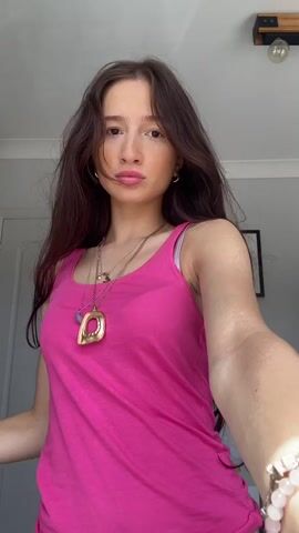 A young woman with long dark hair stands in front of a white door and a white wall. She is wearing a pink tank top, a dark blue skirt with striped accents, and multiple necklaces. She has on bracelets on her left wrist. She is looking at the camera and moving her hands. There is a black tote bag on the floor to her left and a framed picture on the wall to her right.