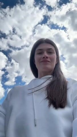 The video shows a woman standing in a mountainous area. She is wearing a white hoodie and has long, dark hair. Behind her, there's a green field with scattered rocks, a small lake, and towering mountains in the distance, some with patches of snow. The sky is partly cloudy. The woman is looking at the camera and seems to be enjoying the view. The camera angle is slightly low, as if she's holding the camera herself.