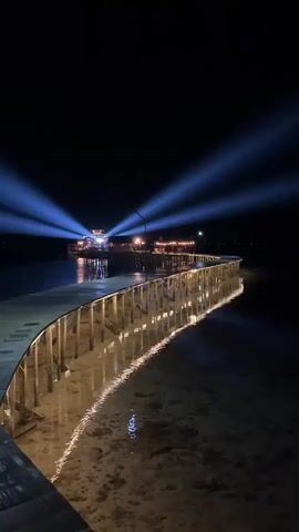 The video shows a night view of a curved wooden pier with lights illuminating its sides. The pier extends over shallow water, revealing the sandy bottom below. In the distance, at the end of the pier, there is a building with multiple lights, and powerful blue spotlights are projected upwards into the dark sky from this area. The scene is dark with the exception of the pier's lights and the spotlights.