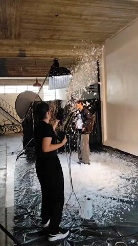 A person in black clothes, holding a black nozzle attached to a cord and a machine, is spraying a white substance that looks like snow onto a man. The man is standing in front of a white backdrop, wearing brown jacket. The floor is covered in the same white substance and a black tarp. There is photography equipment in the background, including a large light with a diffuser, and a bicycle leaning against a wall. The room has a concrete ceiling with visible pipes and light fixtures. There are two bottles on the floor next to the machine.