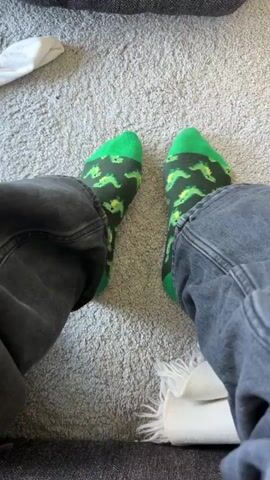 A first-person perspective video shows a person's legs and feet sitting on a gray carpet. The person is wearing dark gray jeans and green socks with a pattern of small green dinosaurs. The feet are slightly moving. In the background, a portion of a gray sofa is visible, along with some white fabric.