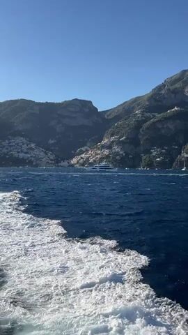 The video shows a view from a boat moving through the water. The camera is angled downwards, capturing the white wake of the boat against the deep blue sea. In the distance, a scenic coastline is visible, characterized by steep, green mountains dotted with white buildings. A large white yacht is anchored in the bay. The sky is clear and blue.