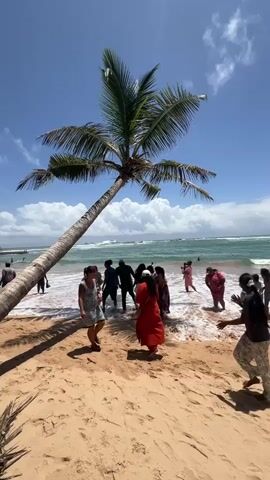 The video shows a beach scene with a large palm tree leaning over the sand. Several people are visible, some walking on the beach and others near the water. One person in the foreground is wearing a light-colored dress and walking towards the right. Another person in a dark shirt and light pants is in motion on the sand. In the background, more people are wading in the ocean, which has waves. The sky is blue with some white clouds. The sand is a light brown color, and the water is greenish-blue. The overall impression is a sunny day at a tropical beach with people enjoying the environment.