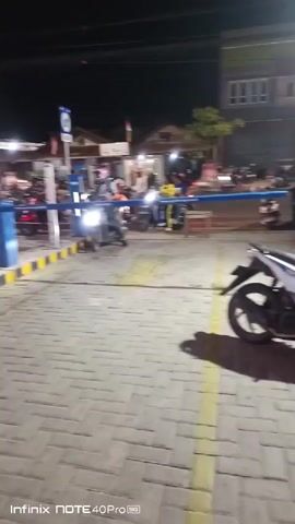 The video shows a nighttime scene of a busy parking lot, likely at a food stall or market, indicated by the presence of multiple motorcycles, people, and illuminated stalls in the background. A blue barrier gate is visible, suggesting an entrance or exit point. A person on a motorcycle is seen passing through the gate. Paved ground with yellow markings is visible. Tires are scattered on the ground. The overall ambiance is lively, with people gathered and vehicles present.