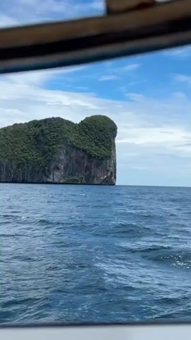 A video depicts a boat ride on a beautiful, clear day. The camera is positioned inside a boat, looking out at a dramatic, lush green island rising from the deep blue ocean. The island is characterized by sheer, rocky cliffs on one side and a dense canopy of trees covering its rounded top. The water is choppy with visible waves, indicating movement and a breezy atmosphere. The sky is a bright blue, dotted with fluffy white clouds. The scene evokes a sense of adventure and natural beauty, suggesting a tropical location. The time of day appears to be daytime, likely late morning or early afternoon, given the bright sunlight. The location is identified as Haad Hin Khom, Thailand. The video captures the impressive scale and ruggedness of the island as the boat sails past.