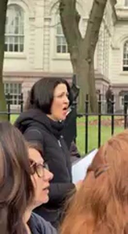 The video captures an outdoor gathering during the daytime, likely in a park or public square. The central subject is a woman with short, dark hair, wearing a black puffer jacket. She is actively speaking or singing, with her mouth open, and holding several white papers in her left hand. Her expression appears earnest and focused.

Partially visible in the foreground are other individuals. To the left, the profile of a woman with dark hair and glasses is seen, seemingly listening intently to the speaker. To the right, the back of a person with reddish-brown hair is visible. In the background, behind the main speaker, another individual wearing sunglasses can be partially observed.

The setting includes trees, which appear bare, suggesting a colder season. A black metal fence separates the immediate foreground from a more distant background. Beyond the fence, a large, light-colored building with multiple windows and classical architectural details stands, consistent with an urban environment like New York, United States, as per the provided context.

The activity seems to be a public address or a group vocal performance. The audio indicates singing or chanting, with the word "tremble" being distinctly audible and repeated, suggesting a protest, vigil, or a communal expression of solidarity or concern. The attire of the participants, including heavy jackets, implies cool or cold weather. There is no discernible text on any objects, clothing, or signs within the frame.