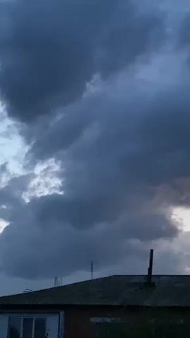 The video shows a dark, ominous sky filled with heavy storm clouds. The clouds are a deep blue-gray, with some lighter patches where the sunlight is trying to break through. The camera is pointed upwards, capturing the dramatic sky above a rooftop. The rooftop appears to be part of a brick building, with a dark, sloped roof. There are a few objects on the roof, including a chimney, an antenna, and what looks like a satellite dish. The overall impression is one of an approaching storm, with the weather creating a somber and dramatic atmosphere.