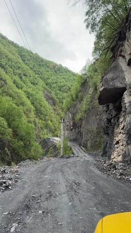 The video shows a dirt road winding through a steep, forested mountain valley. The road is narrow and appears to be in poor condition, with loose gravel and dirt. The mountainsides are covered in lush green trees and vegetation. The sky is overcast, and the lighting suggests it may be daytime, possibly late afternoon or early morning. There is a power pole with wires running along the side of the road. A yellow object, possibly the corner of a vehicle, is visible in the bottom right corner of the frame, suggesting the video is being recorded from a moving vehicle. The road leads towards a bridge or elevated section in the distance. The overall impression is one of a remote, rugged, and possibly challenging natural environment.